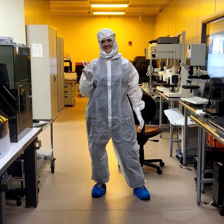 Gowned in a full cleanroom suit, hood and overshoes, standing in the fabrication bay between benches of microscopes and process equipment.