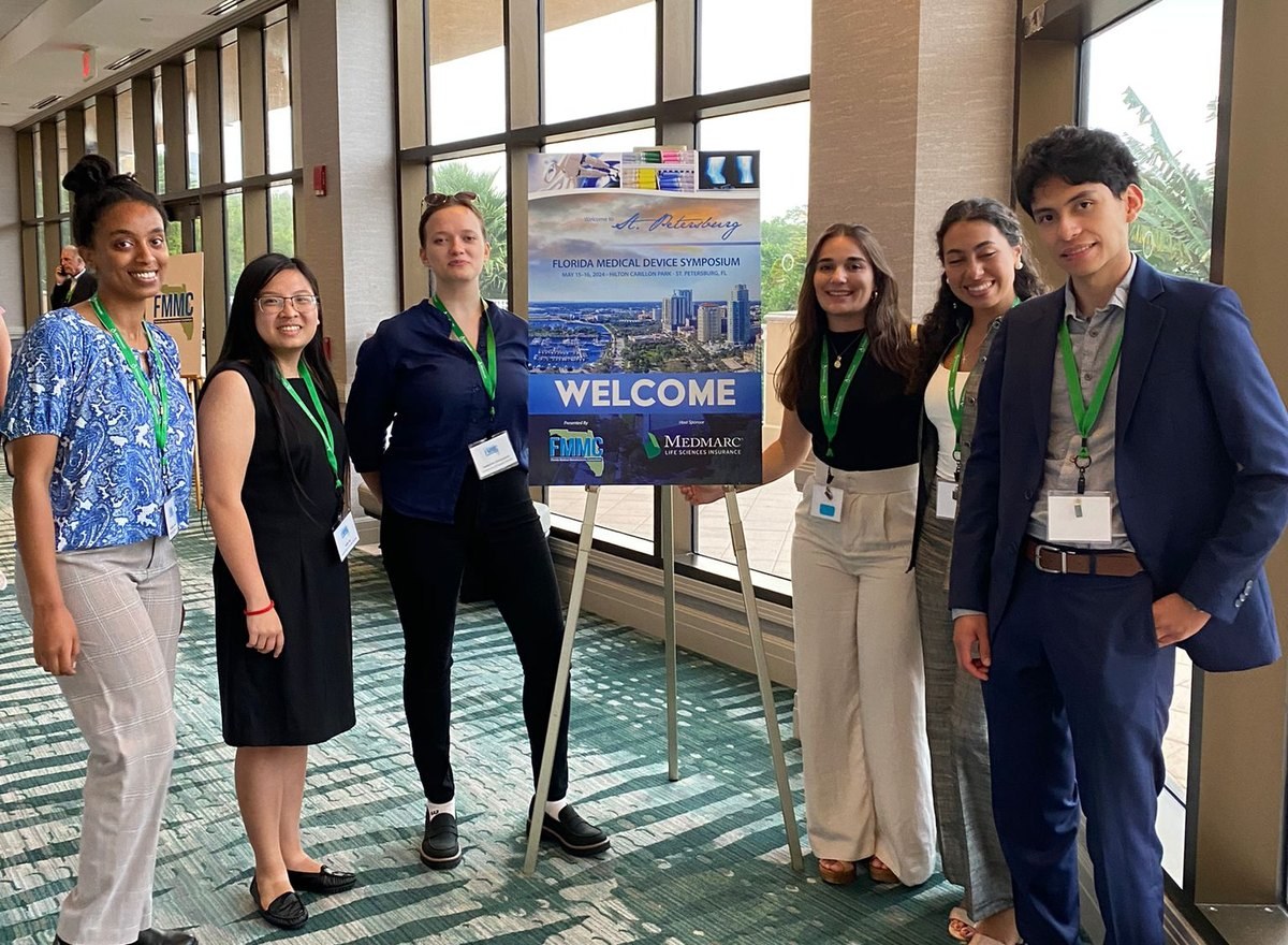 Six of us standing beside the welcome board at the Florida Medical Device Symposium in St. Petersburg, all wearing lanyards.
