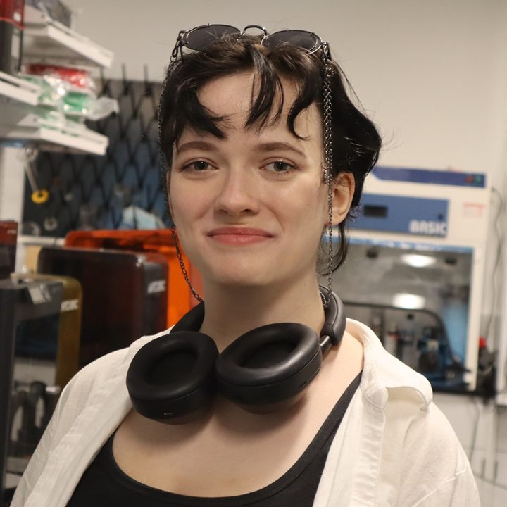 Nadia Korostyleva in the lab, headphones round the neck and sunglasses pushed up into short dark hair, with a fume hood and printers behind.