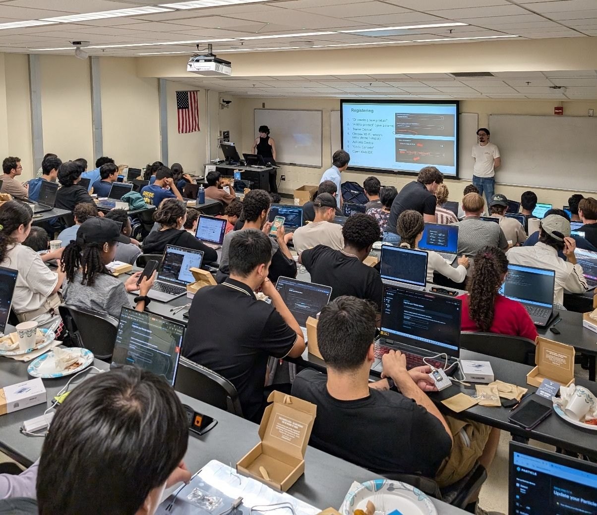 A packed lecture room seen from the back: every seat taken, laptops open along the benches with kit boxes beside them, and a slide projected at the front.